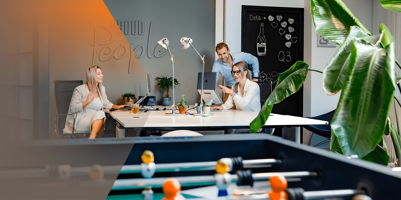 Vooraanzicht van twee dames en een heer aan een tafel. Een van de dames wijst iets aan op de laptop in haar hand en de heer en andere dame lachen ernaar.